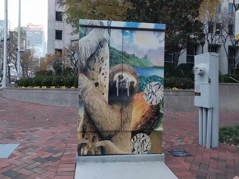 A painted utility box in an urban plaza features a realistic sloth clinging to a tree, with a scenic mountain and lake…