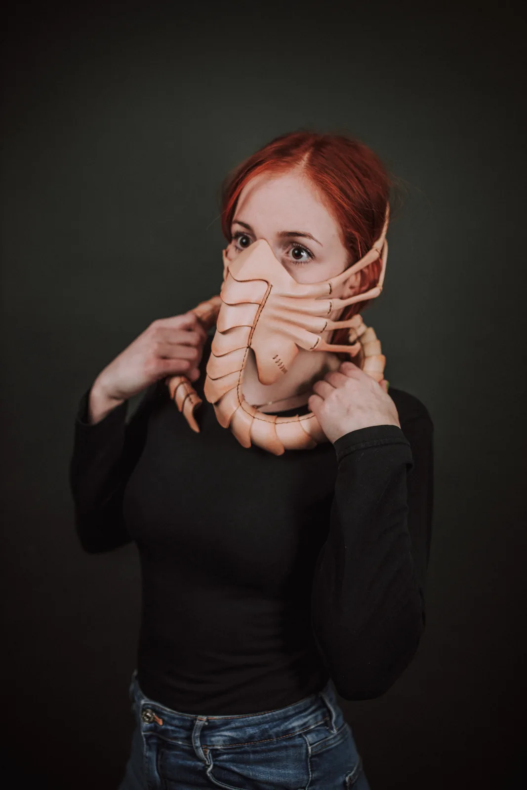 A woman with red hair, wearing a black long-sleeved shirt and jeans, holds a large, tan-colored, segmented mask resembling a…