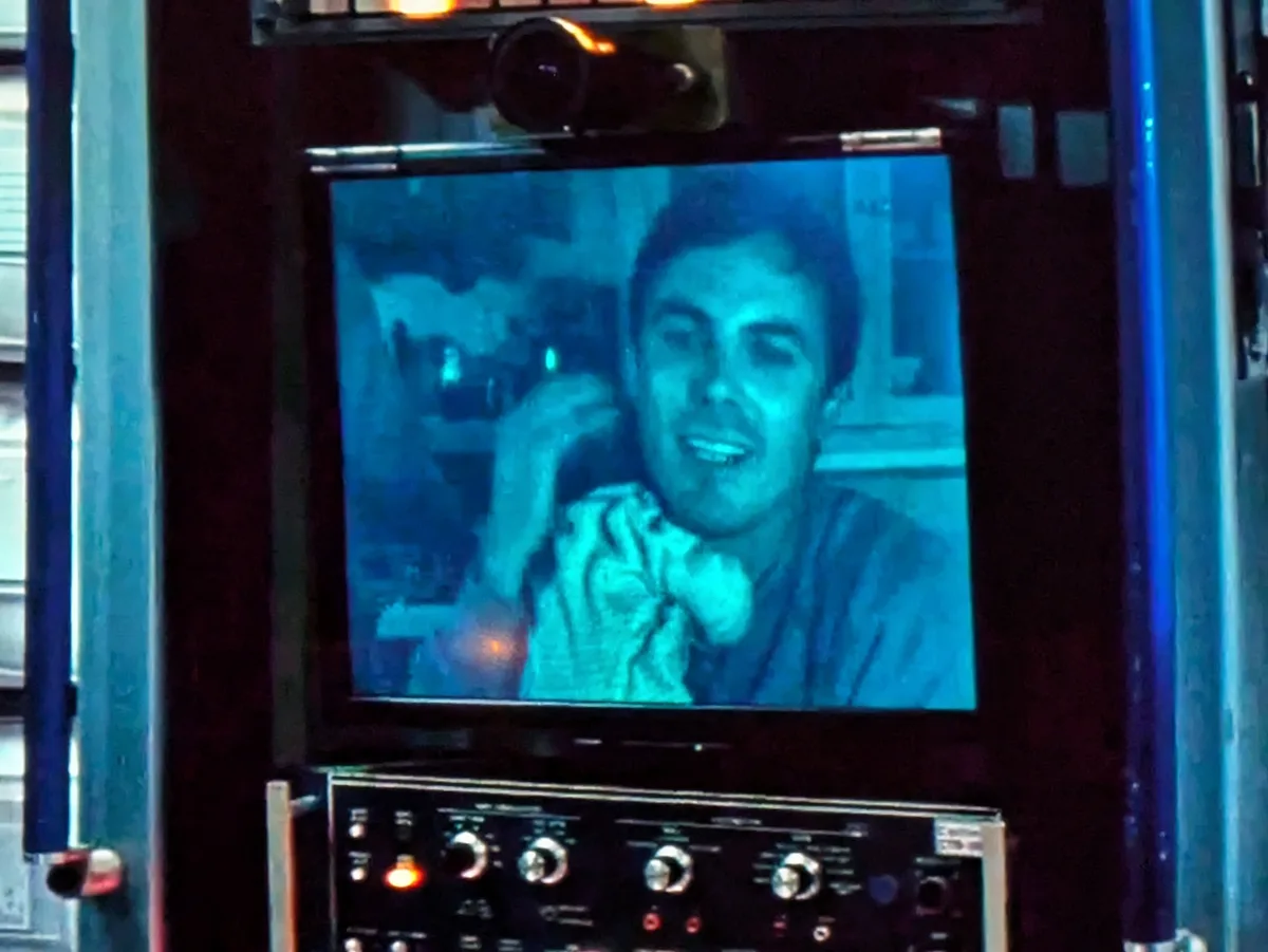 A grainy, blue-tinted security camera feed shows a smiling man holding a phone to his ear, displayed on a monitor mounted…