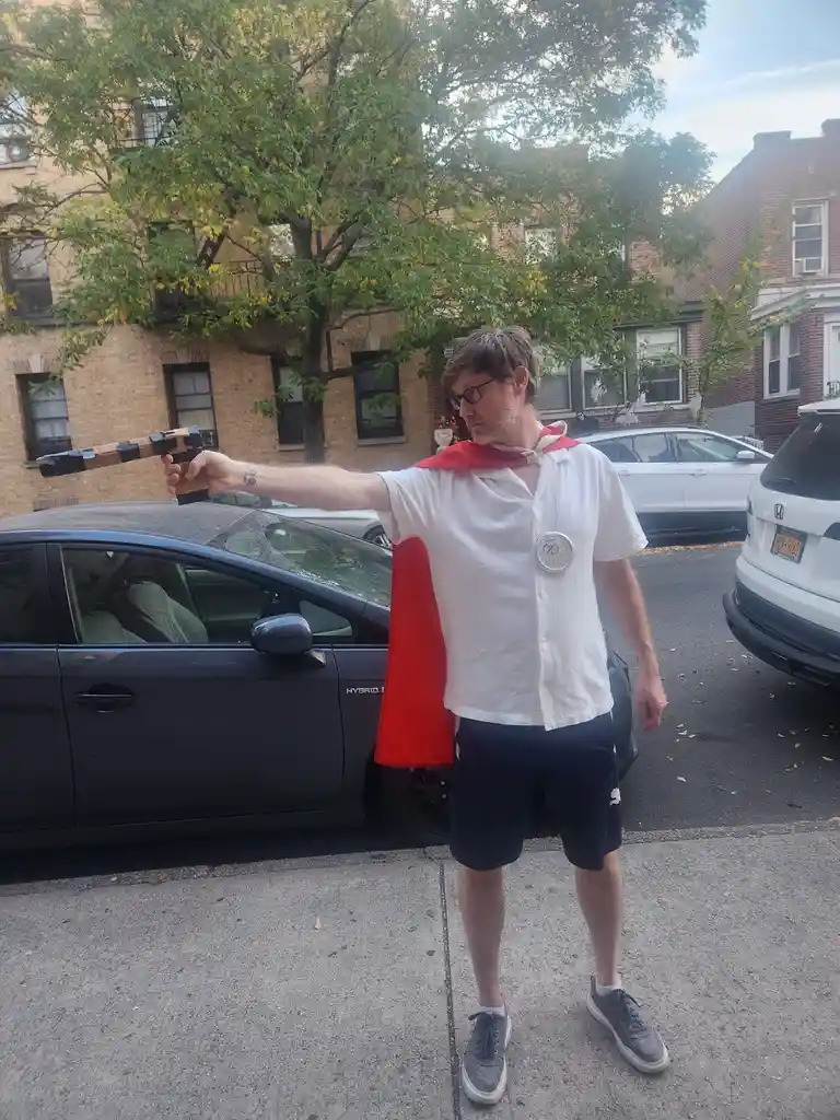 A man wearing a red cape and a white shirt with a silver medal around his neck stands on a sidewalk in front of parked cars…
