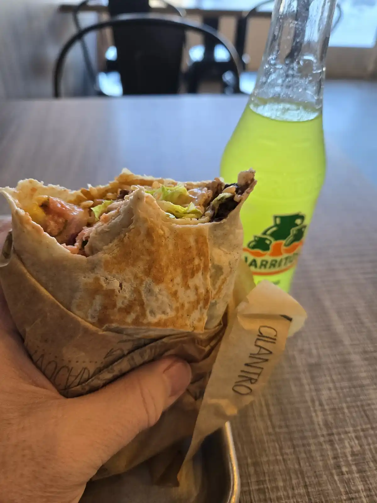 A hand holds a partially eaten burrito wrapped in brown paper with 'CILANTRO' printed on it, next to a green bottle of…