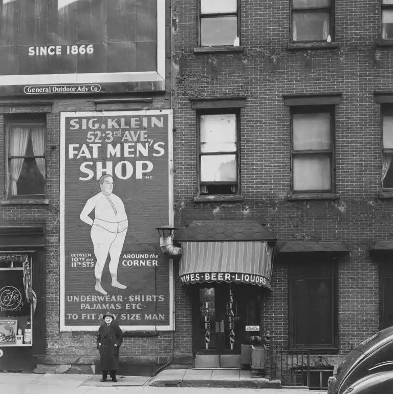 A vintage black-and-white photo shows a large billboard for 'Sig. Klein, Fat Men's Shop' advertising underwear and pajamas…