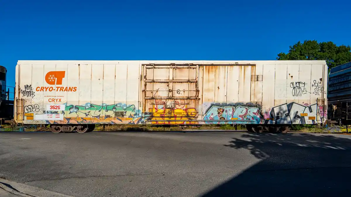 A weathered white Cryo-Trans freight train car, marked with the number 3525, is covered in colorful graffiti under a clear…