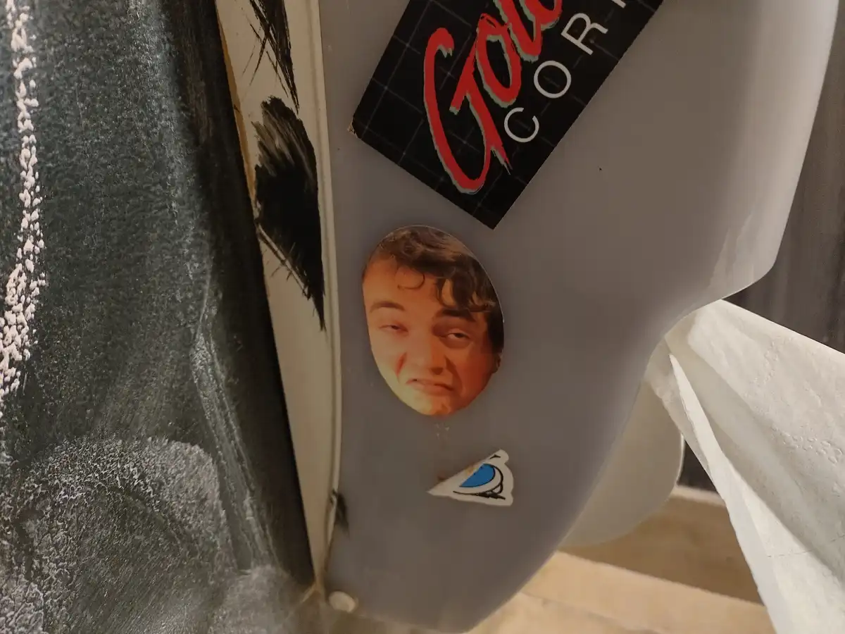 A close-up photo shows a grey toilet paper dispenser with a circular cutout revealing a man's face looking slightly annoyed…