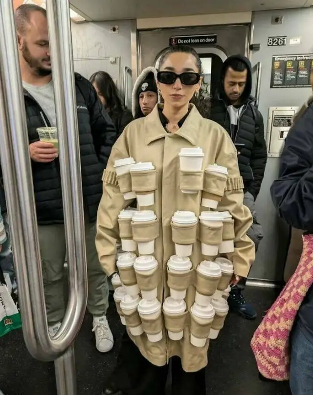 A person on a subway train wears a beige coat with multiple pockets holding white disposable coffee cups. The individual…