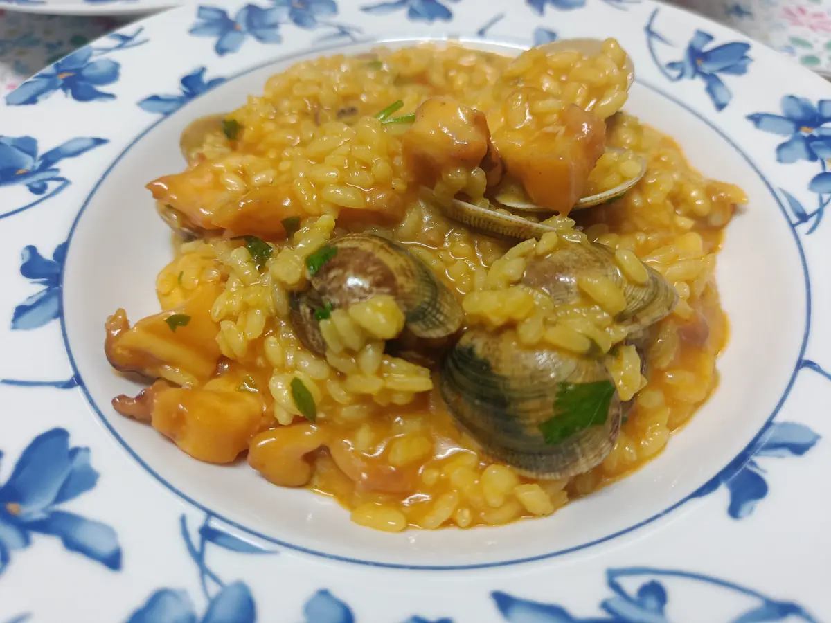 A close-up photo shows a plate of seafood risotto with clams, shrimp, and a creamy sauce, served on a white plate with blue…