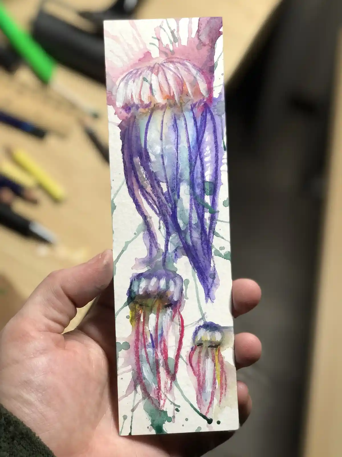 A hand holds up a small rectangular watercolor painting depicting three stylized jellyfish in purple, pink, and red hues…