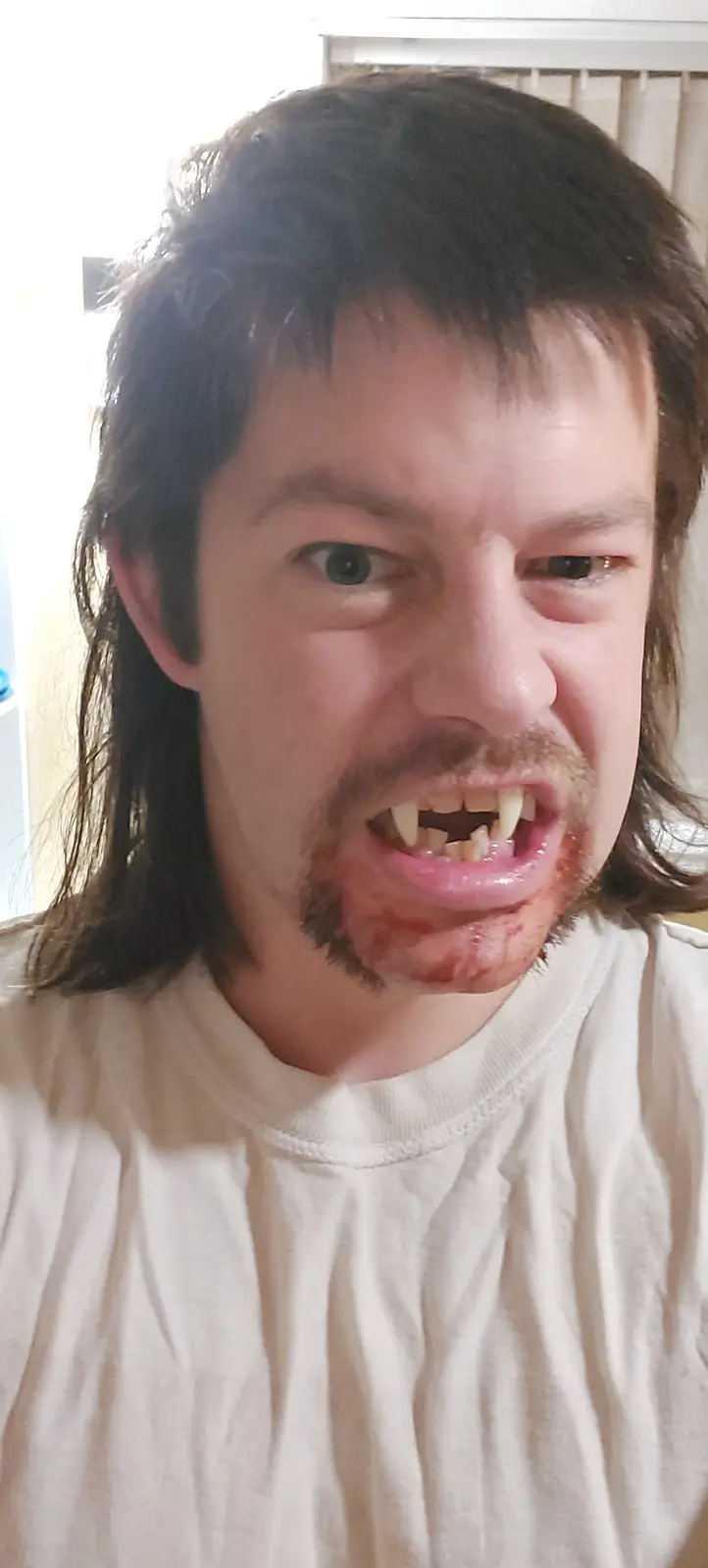 A man with long dark hair and a goatee stares intensely at the camera with his mouth open, revealing fake fangs and what…