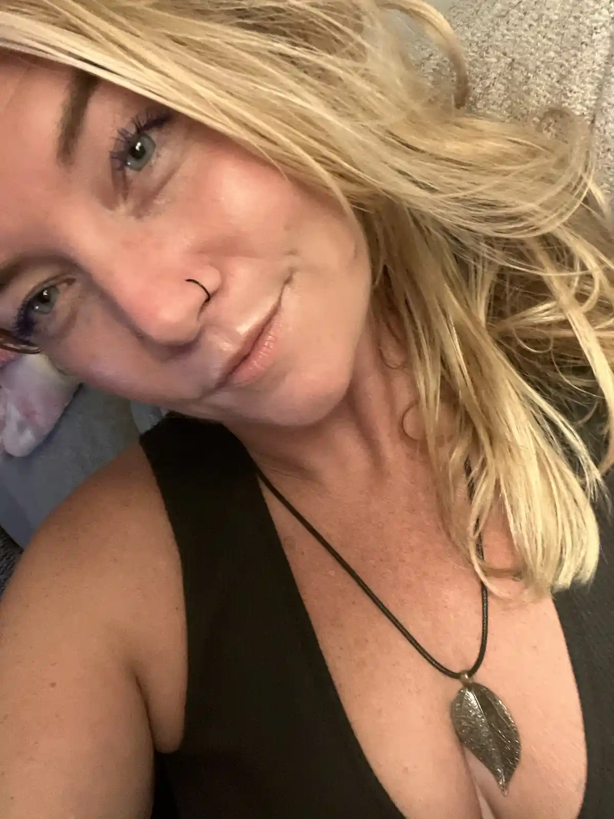 A selfie of a woman with blonde hair and green eyes, wearing a black top and a leaf-shaped pendant necklace, looking at the…