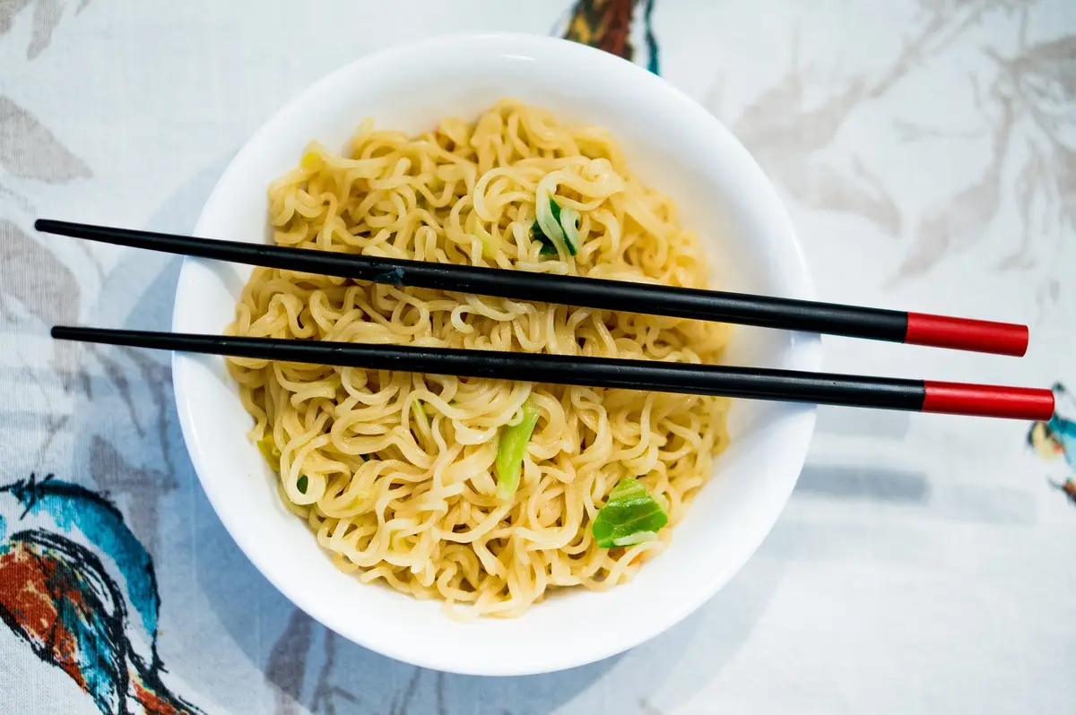 A top-down photo of a white bowl filled with cooked ramen noodles and chopped green onions, with black chopsticks with red…