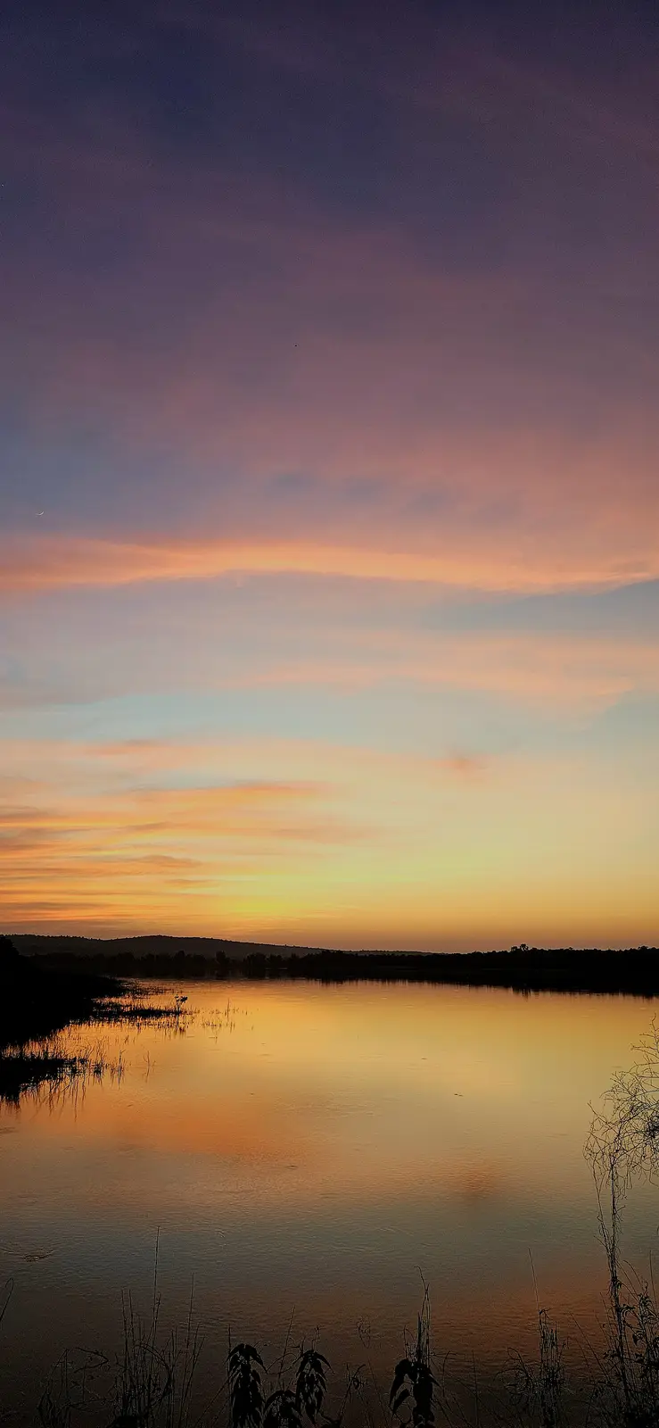 A serene sunset over a calm body of water, with a gradient sky transitioning from deep purple to orange and yellow near the…