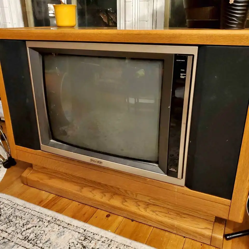 A vintage Sharp CRT television set, housed in a wooden console with large black speakers on either side, sits on a hardwood…