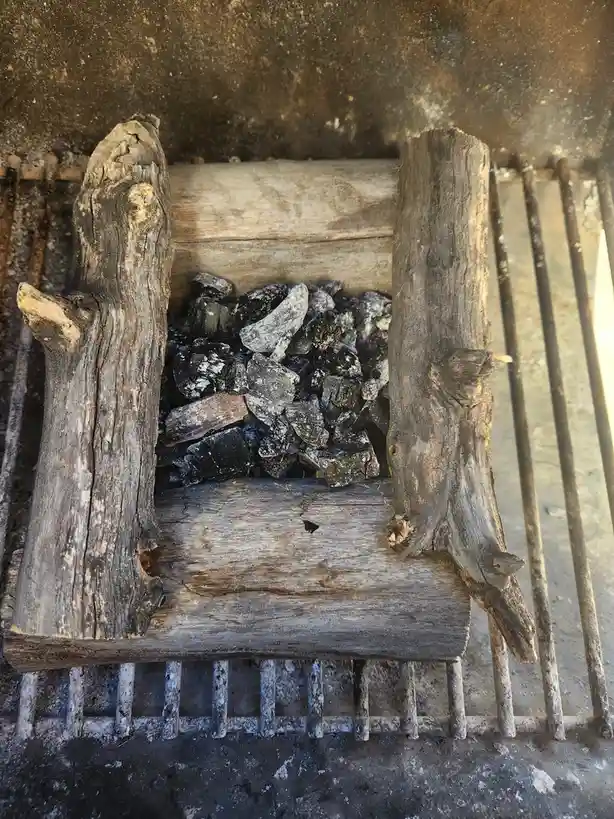 a close-up photo shows a grill with charcoal briquettes inside a wooden structure shaped like a chair, framed by two upright…