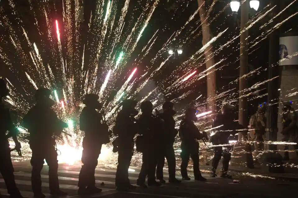 A nighttime photo shows silhouettes of police officers in riot gear standing on a street as bright red and green fireworks…