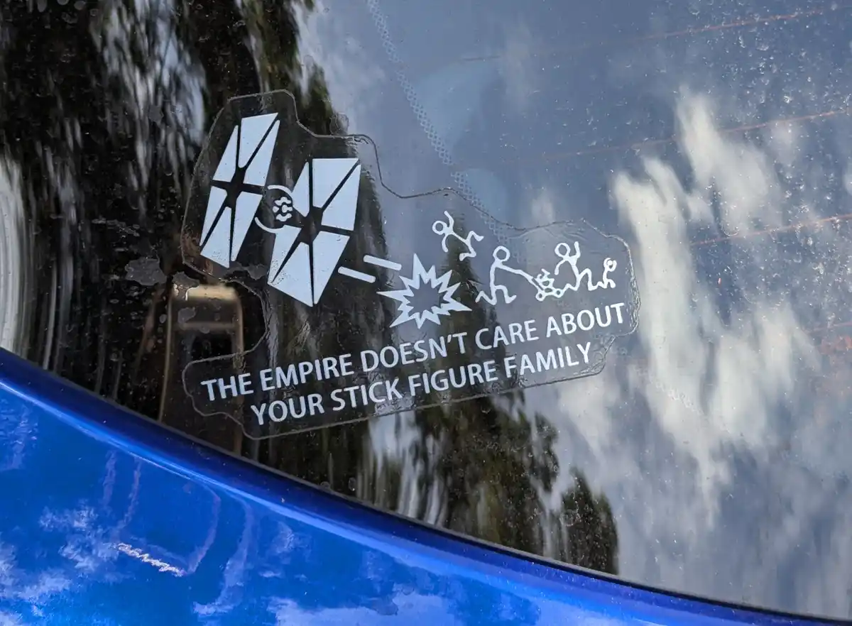 A close-up of a car window sticker showing a Star Wars Imperial emblem firing at stick-figure family members, with the text…