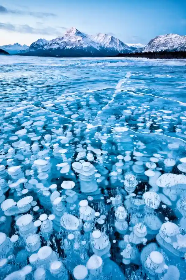 A stunning photograph of a frozen lake covered in numerous ice bubbles, with snow-capped mountains in the background under a…