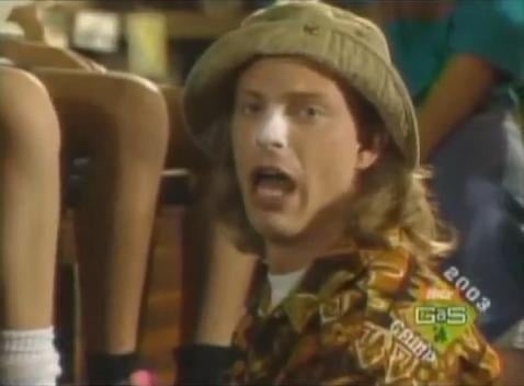 A man with long hair and a beige bucket hat wears a colorful patterned shirt and stares with an open mouth in a scene from…