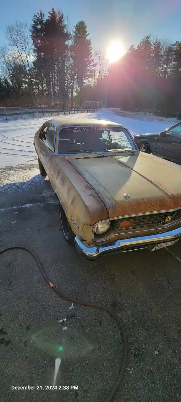 A rusty, weathered vintage car with a Ferrari emblem on its grille is parked in a snowy driveway under a bright sun. A hose…