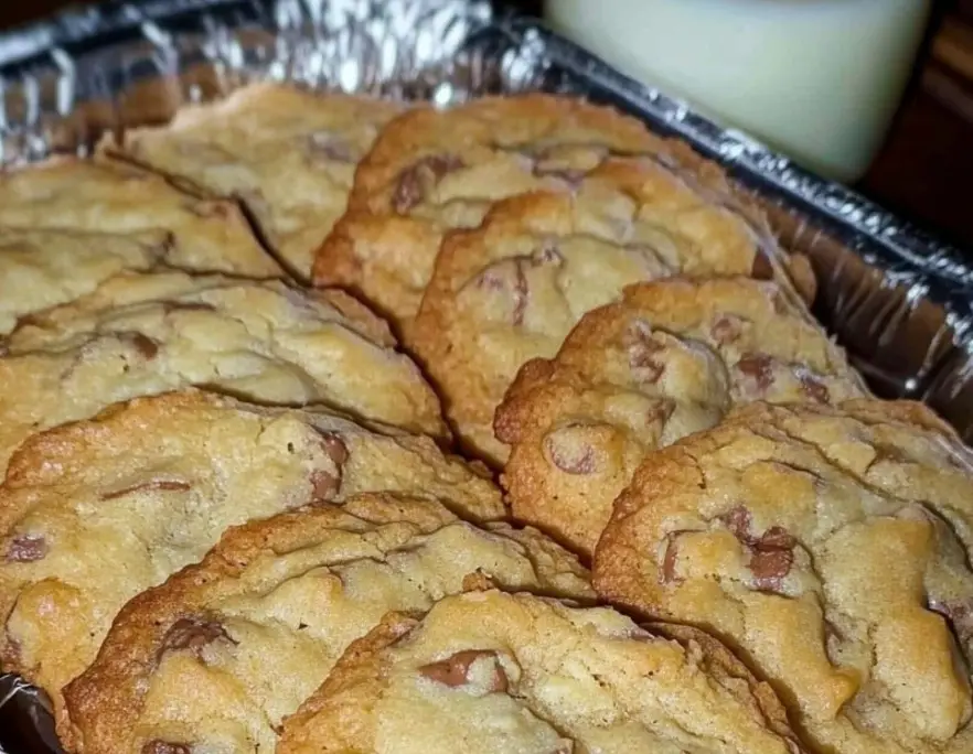 A close-up photo shows a foil-lined baking pan filled with golden-brown chocolate chip cookies, with a glass of milk visible…