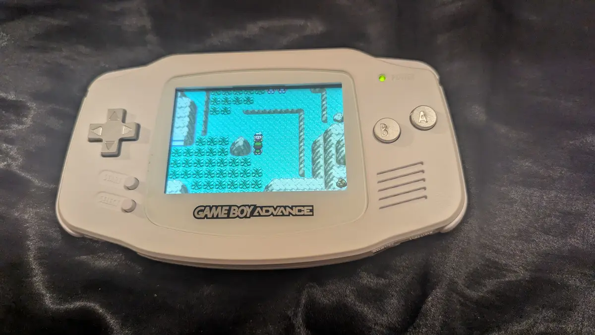 A white Game Boy Advance console rests on a dark fabric surface, its screen displaying a pixelated Pokémon game scene with a…