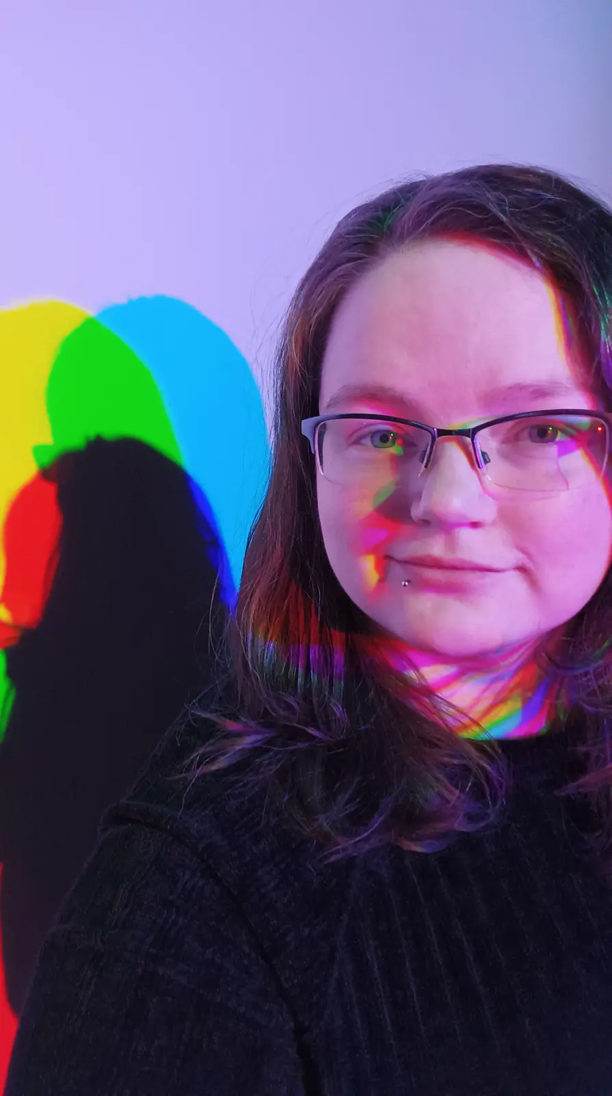 A person with glasses and a lip piercing looks at the camera while colorful light projections (yellow, green, blue, red)…