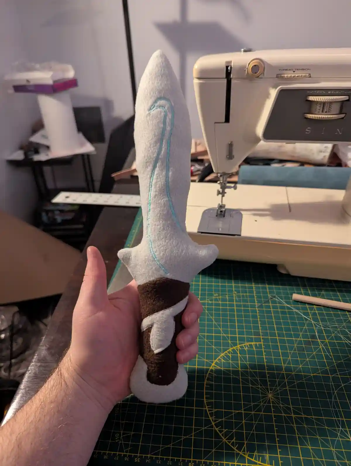 A hand holds a handmade felt sword with a brown and white handle and a light gray blade with a light blue stitched line, in…