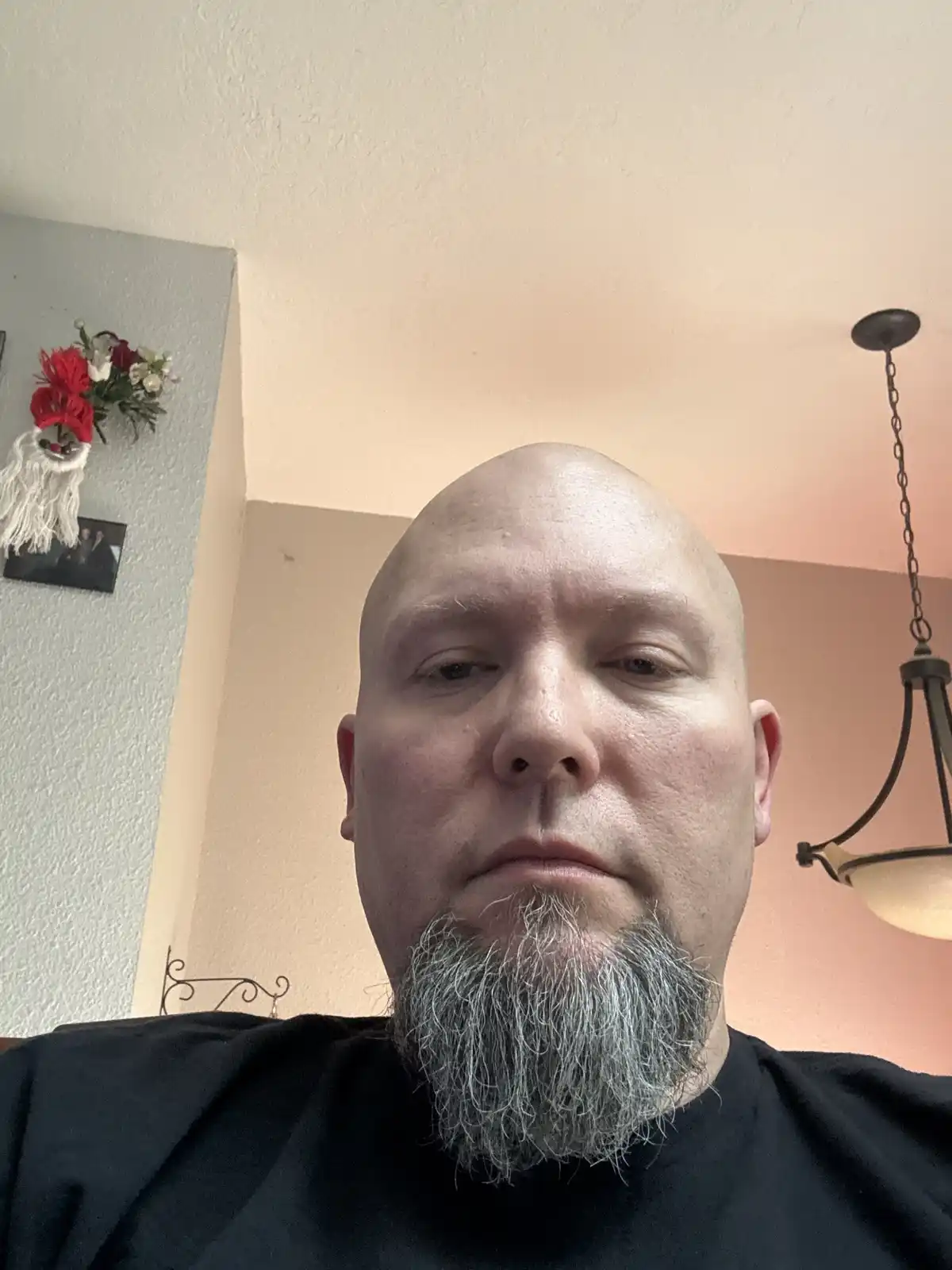 A bald man with a full, gray goatee stares directly at the camera with a neutral expression. He wears a black shirt and is…