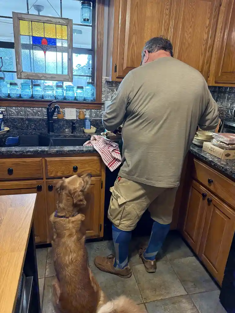 A man in a kitchen, viewed from behind, is standing at a counter preparing food while a golden retriever sits on the floor…