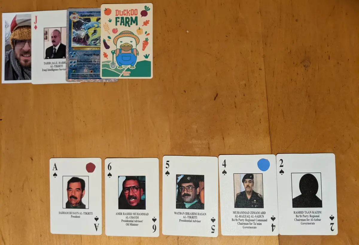 A wooden table displays a row of custom playing cards featuring photos and titles of Iraqi officials, including Saddam…