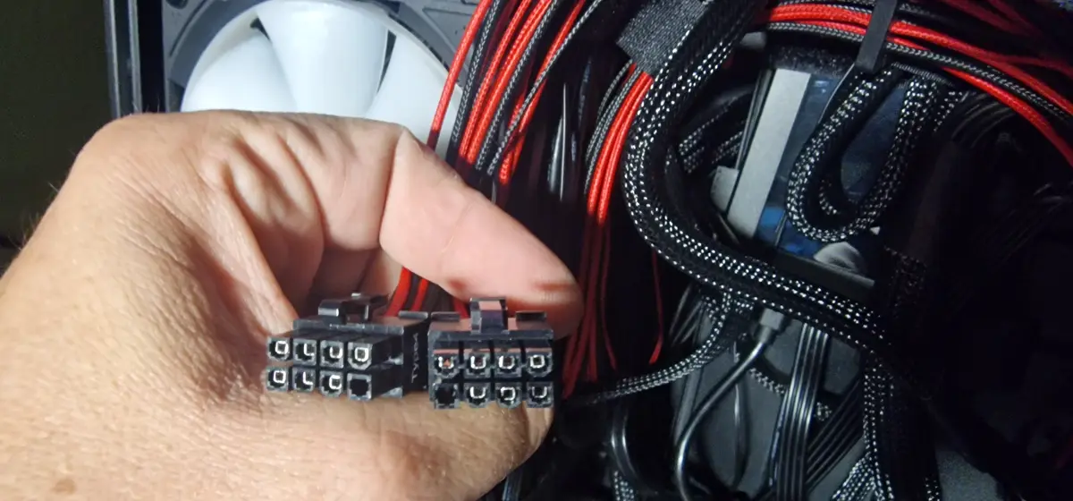 A hand holds two small black 24-pin ATX power connectors against the backdrop of a computer case interior, which features…