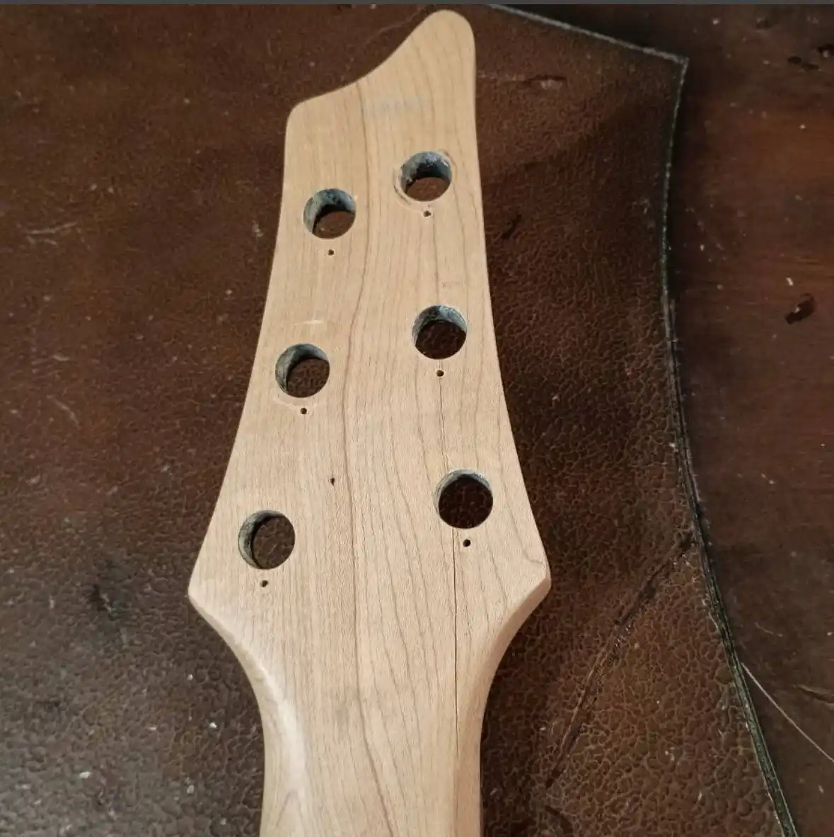 A close-up photo shows the headstock of a light-colored wooden bass guitar, featuring six tuning peg holes arranged in two…