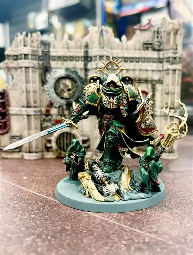 A painted miniature of a green-armored, hooded warrior with a sword and ornate staff stands on a base with two smaller…