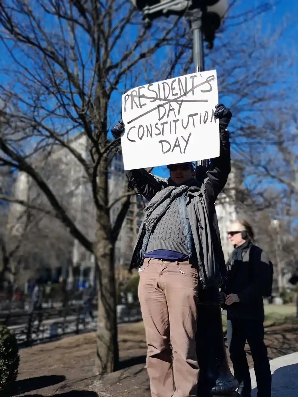 A person in a winter jacket and scarf holds a white sign outdoors with bare trees and a blue sky behind them. The sign has…