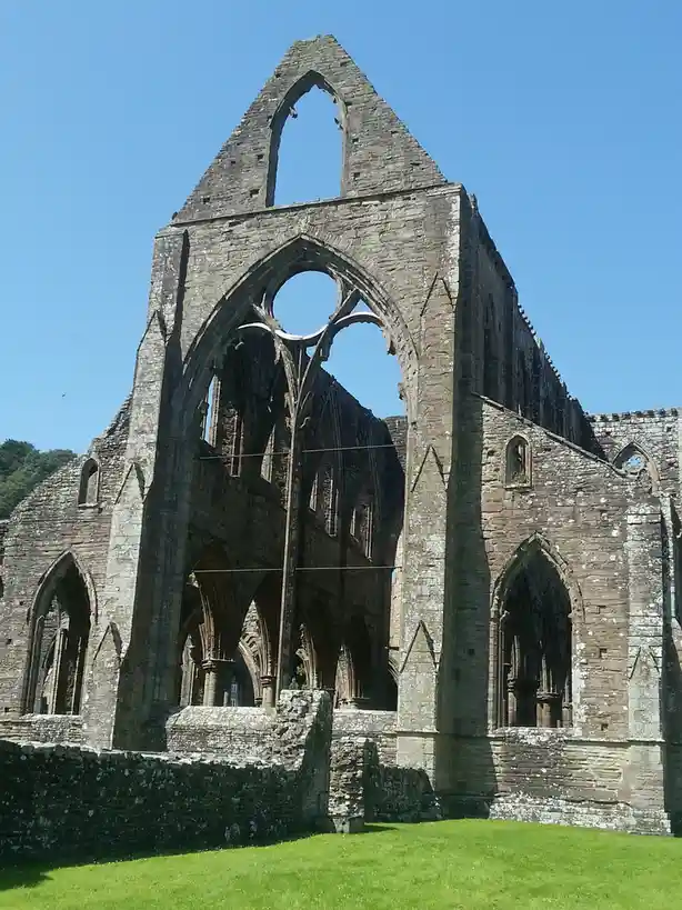 The image shows the ruins of a Gothic-style stone abbey under a clear blue sky, with a vibrant green lawn in the foreground…