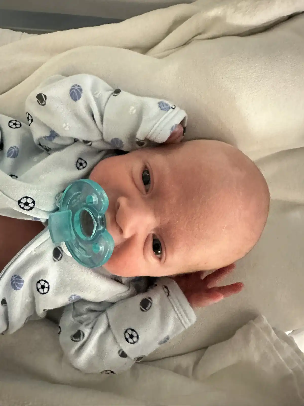 A newborn baby with a pacifier in its mouth lies on a white blanket, wearing a light-colored onesie with black and blue…