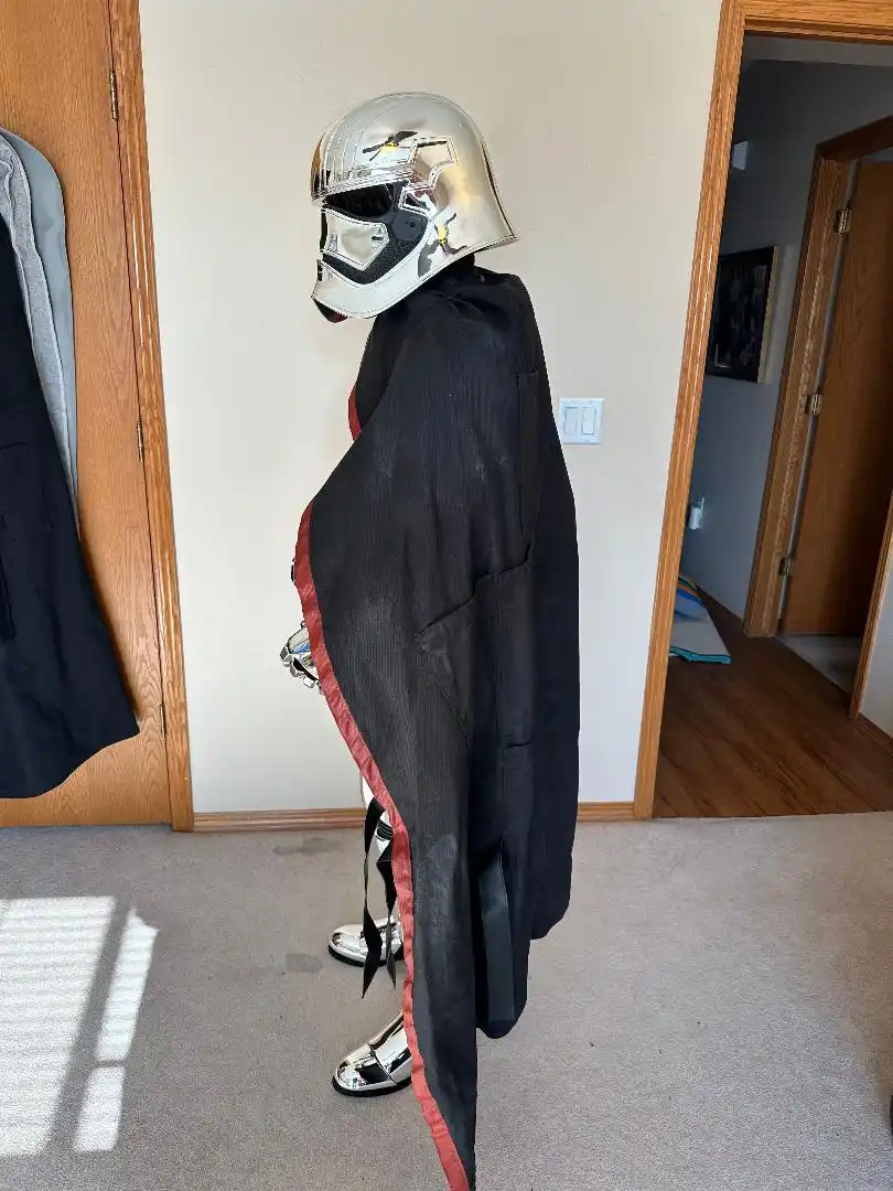 A person in a silver stormtrooper helmet and black cape with red trim stands in profile in a carpeted room, facing away from…