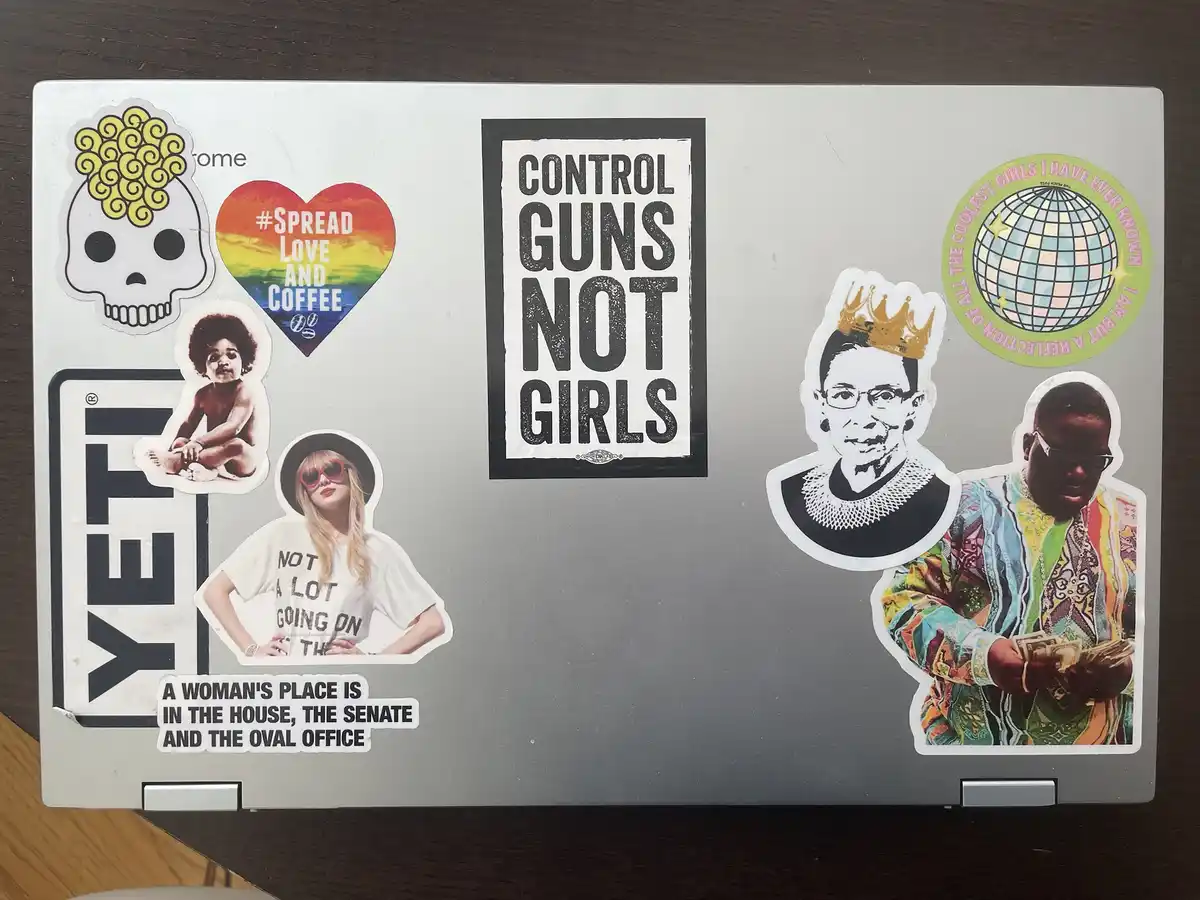 A silver laptop lid covered in various stickers, including a skull with yellow hair, a rainbow heart with '#SPREAD LOVE AND…