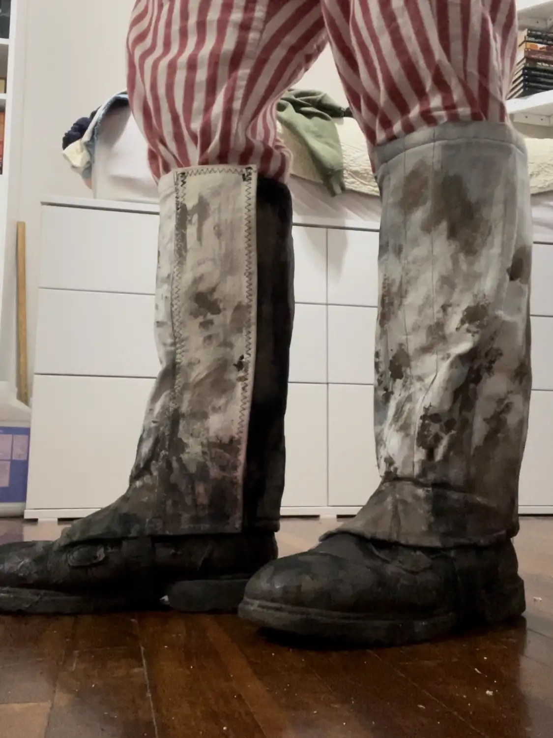 A person wearing red and white striped pajama pants stands indoors on a wooden floor, their boots and lower legs covered in…