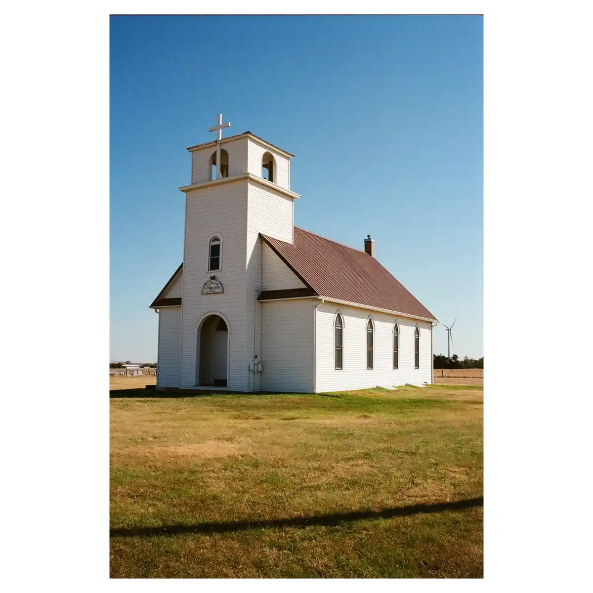 A white wooden church with a steeple and cross stands alone in a grassy field under a clear blue sky, with a wind turbine…