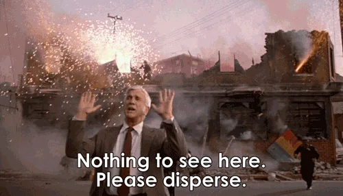 A man in a suit and tie stands in front of a burning, rubble-strewn street with explosions and debris flying around him…