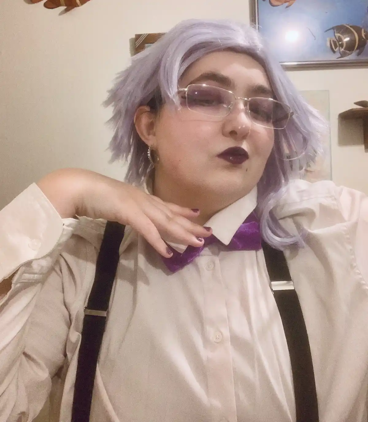 A person with short, messy lavender hair, wearing clear-framed glasses, dark lipstick, and a white button-up shirt with…