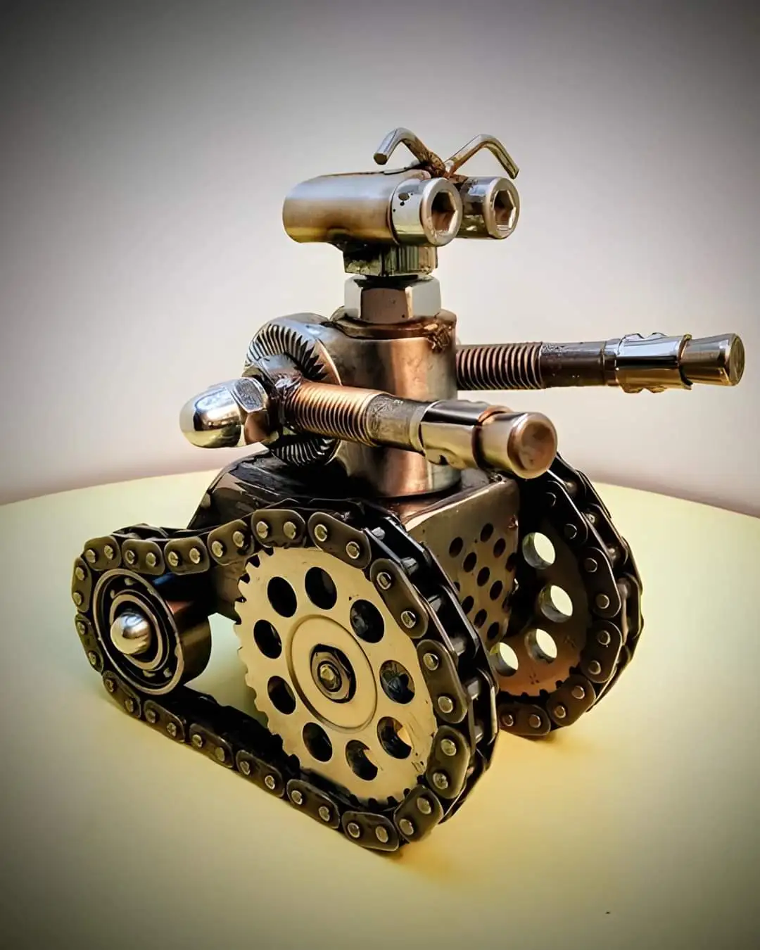 A handmade robot sculpture crafted from metal parts like chains, bolts, gears, and pipes, resembling a tank with a head made…
