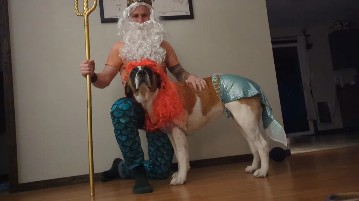 A man dressed as a king with a white wig, beard, and blue scale leggings kneels while holding a golden trident. Beside him…