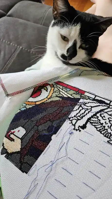 A black and white cat with a sleepy expression rests its head on a partially completed cross-stitch project depicting a…