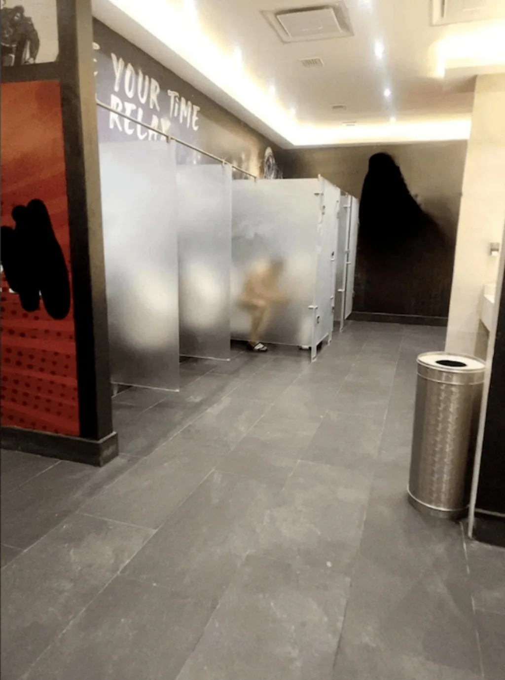 A blurry photo of a public restroom with frosted glass stalls, a person motion-blurred inside one stall, and a large dark…