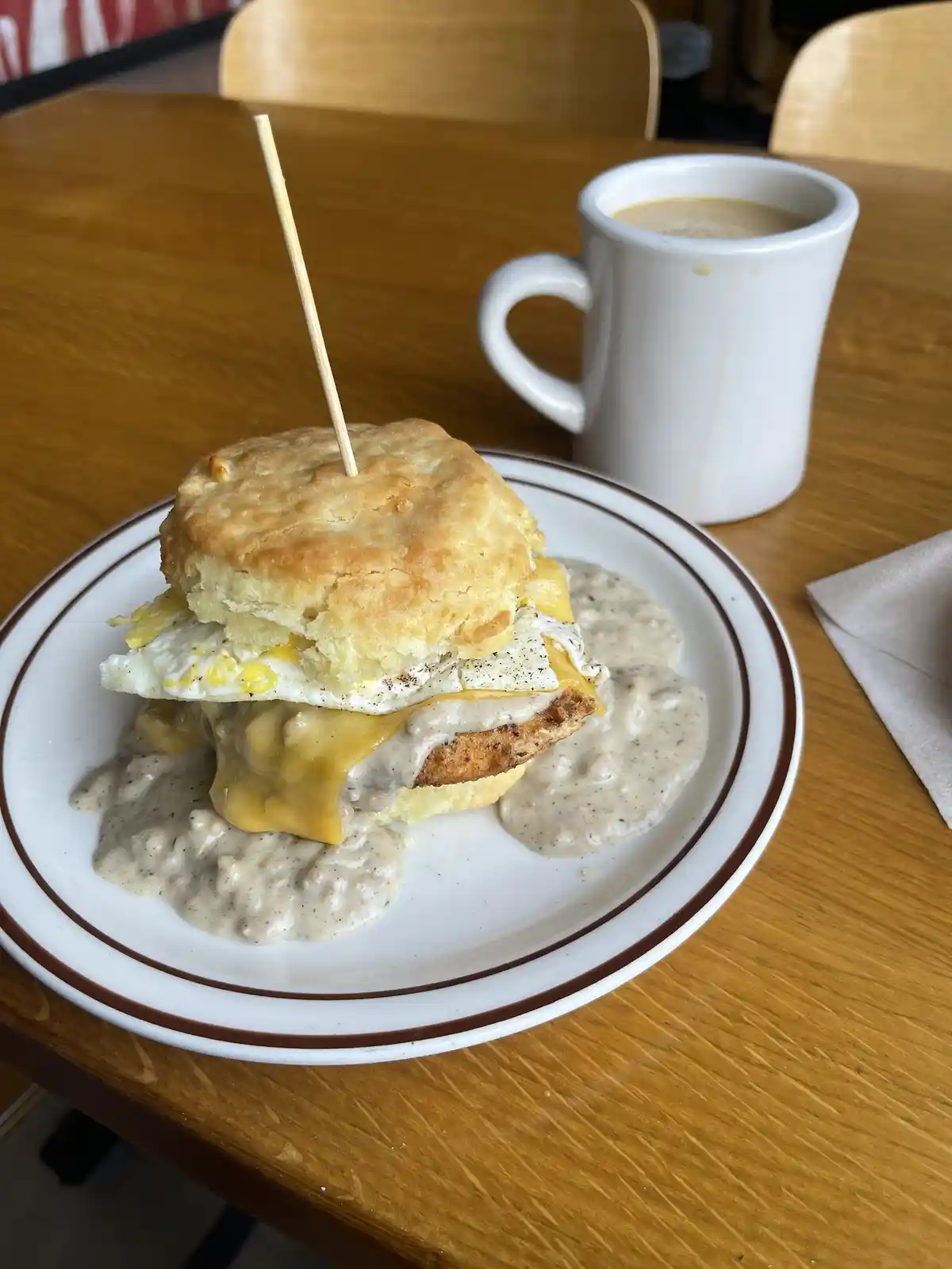 A close-up photo shows a breakfast biscuit sandwich on a white plate with brown trim, sitting on a wooden table. The…