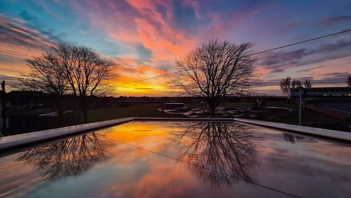 A serene sunset with vibrant orange, pink, and blue clouds reflected in a calm pool of water, framed by the silhouettes of…
