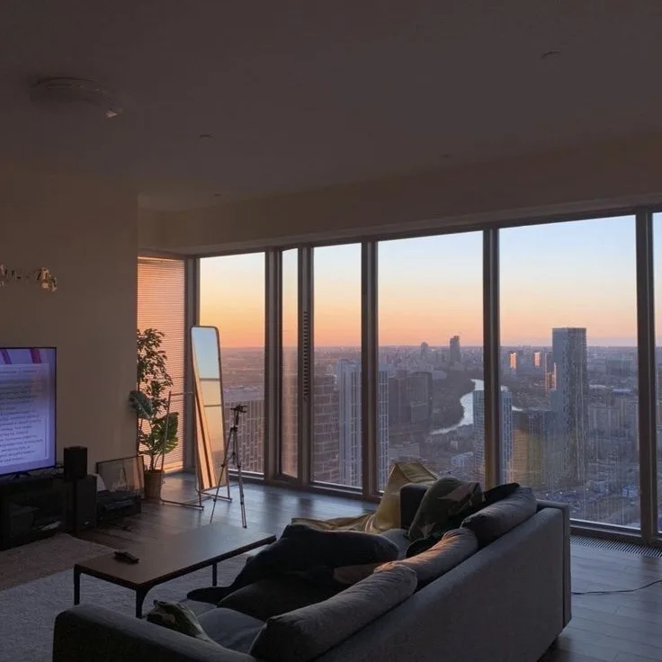 A cozy, modern living room with a large sofa facing floor-to-ceiling windows showing a stunning city sunset. A TV is on in…