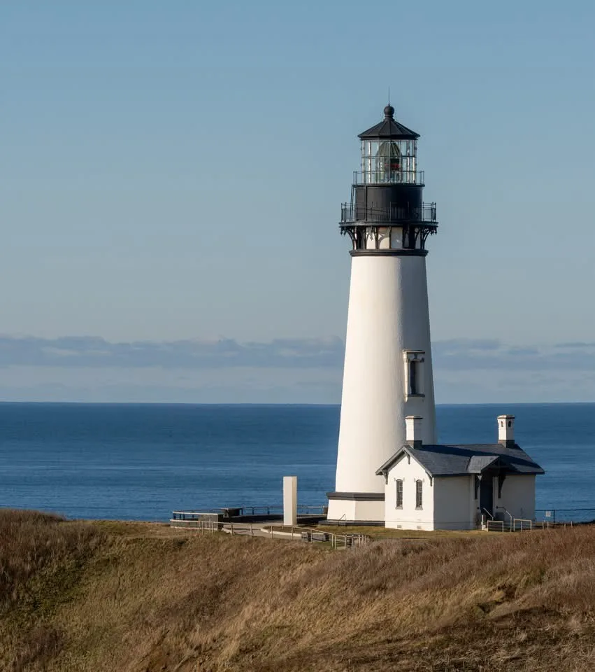 A tall white lighthouse stands on a grassy cliff overlooking the ocean under a clear blue sky. The lighthouse has a black…