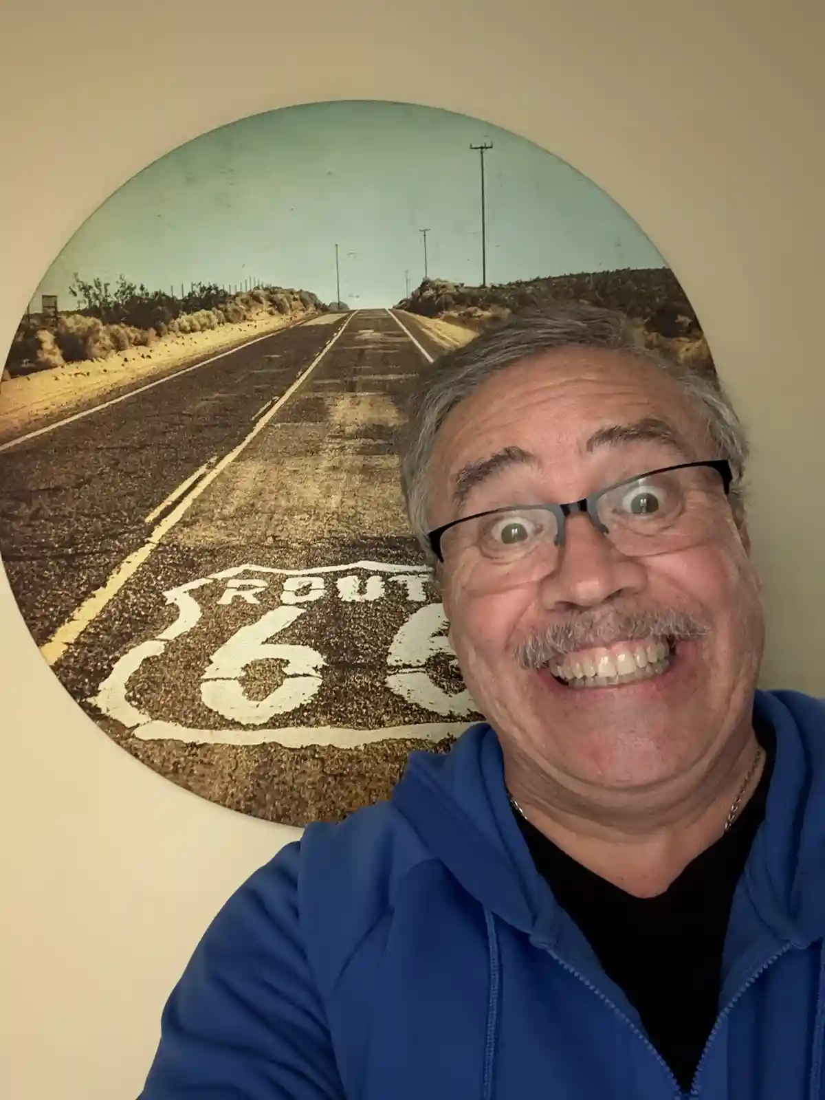 A man with glasses and a mustache makes a wide-eyed, excited expression while taking a selfie in front of a circular wall…